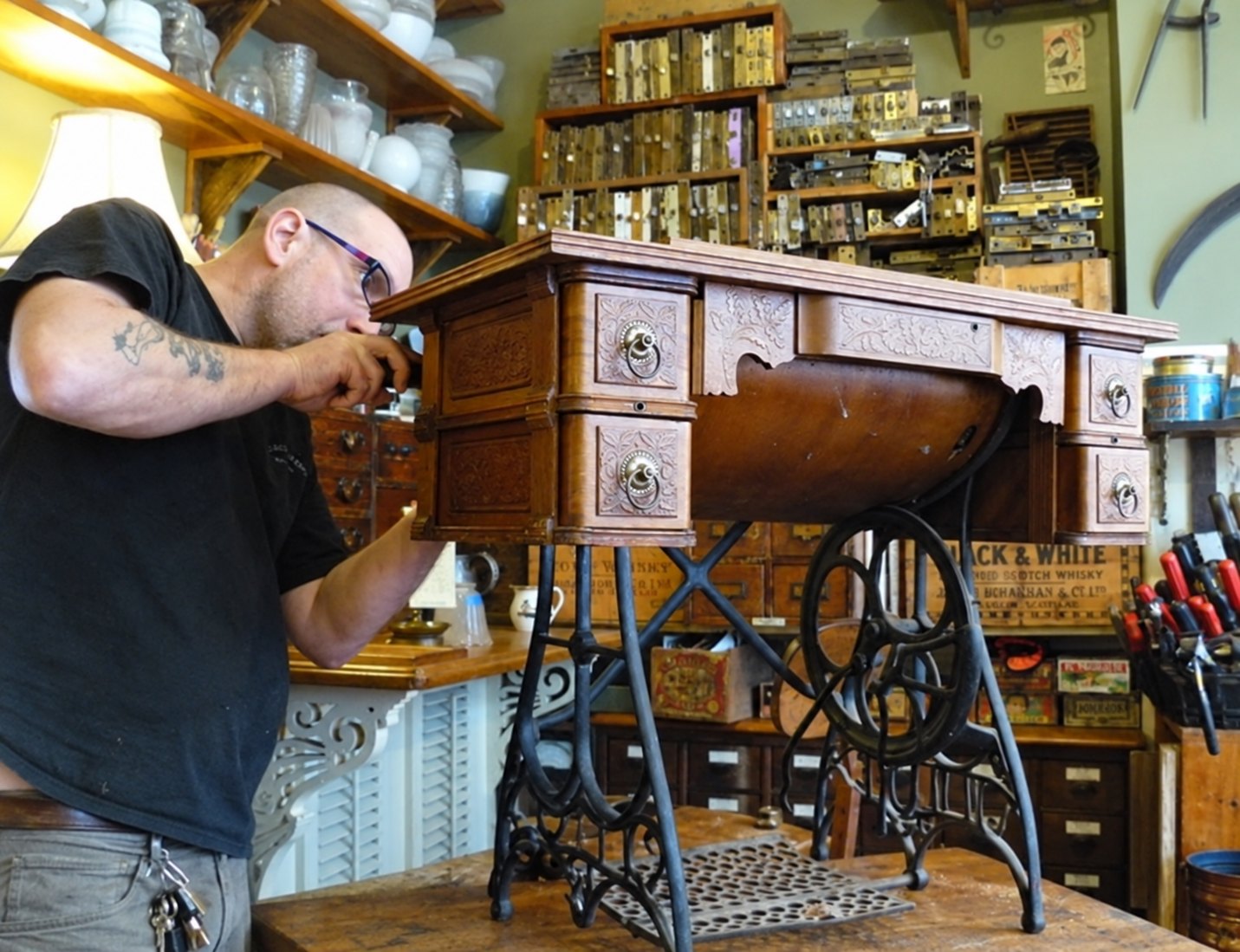 Seth leans over an antique treadle sewing machine table on the workbench, adjusting the carved upper corner of the cabinet above its cast-iron treadle and drive wheel.