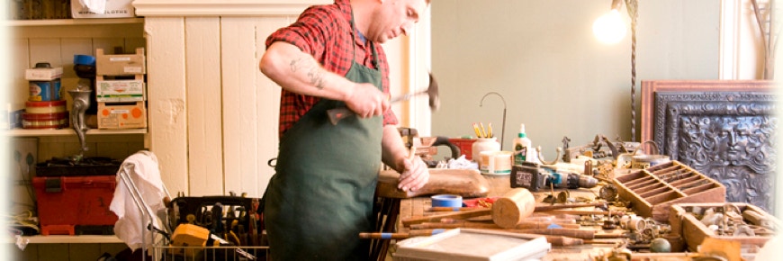 Seth in a green work apron at his workbench, tapping a chisel with a hammer, surrounded by tools and pieces in for repair.